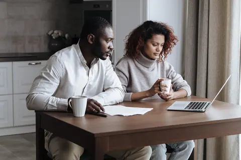 Two adults sit side by side at a kitchen table, looking at a laptop and papers together.