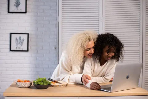 A woman and a young girl look at a laptop together at a kitchen counter.
